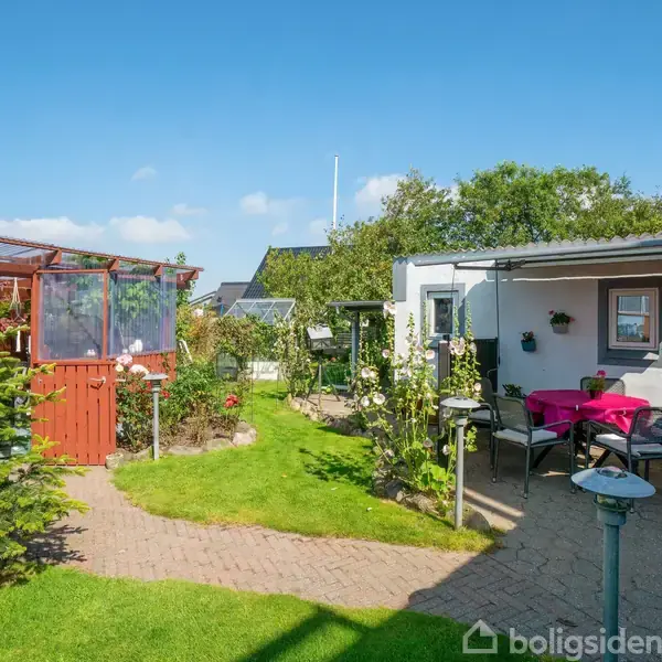 En lille have med grøn græsplæne omgivet af buske. En rød træbygning står til venstre; mod højre er der en terrasse med et bord og lyserødt dug under en åben himmel.