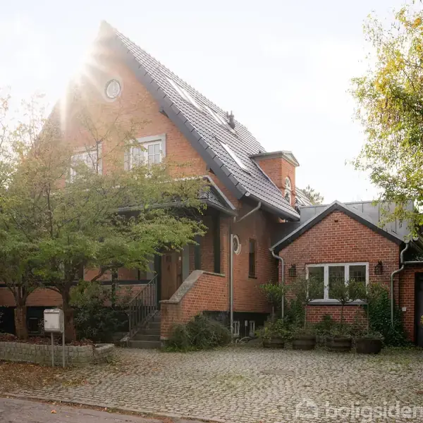 En rød murstensvilla med sadeltag står i sollys. Der er en brolagt indkørsel foran huset, omgivet af træer og buske, hvilket skaber et hyggeligt miljø.