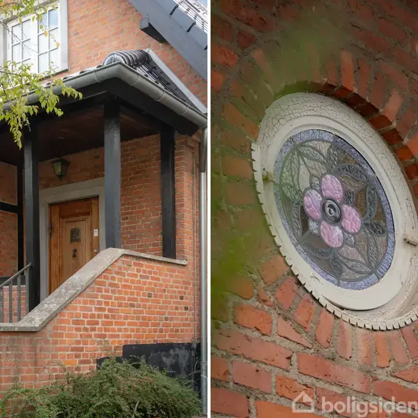 Siddet af en rød murstensbygning viser en hoveddør med trappe og et rundt vindue med farvet glas i en blomstermotivramme. Grønne blade omkranser delen af huset.