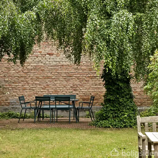 Et sort havebord med seks stole står på en rødlige terrasse omgivet af grønne træer og buske. I baggrunden ses en murstensvæg, og forgrunden har en træbordkant.