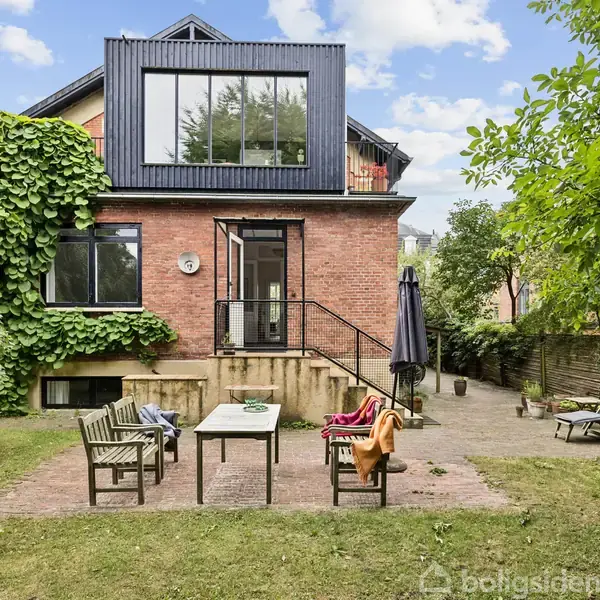 Murstenshus med moderne tilbygning. I forgrunden står havemøbler på terrasse omgivet af græsplæne og træer. En trappe fører til husets hoveddør.