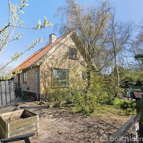 Et gulstenshus med rødt tag står omgivet af træer og buske i en have. En træbeholder og træhegn ses i forgrunden.