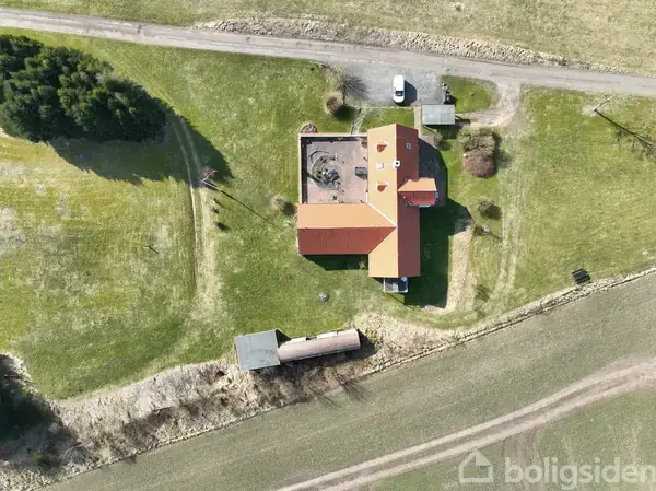 En stor villa med rødt tag set fra oven, omgivet af grønne marker og en grusvej. Flere træer står tæt ved huset, og en bil holder parkeret i indkørslen.