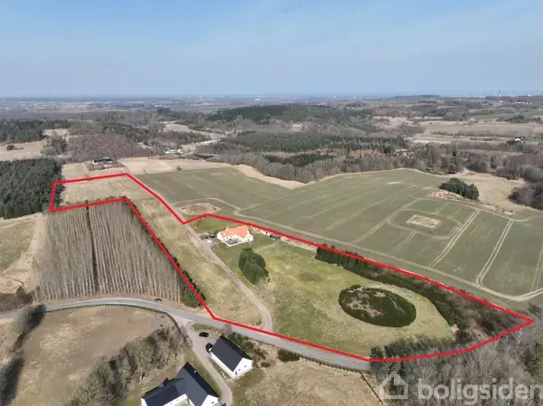 En landejendom med rødt tag ligger centralt omgivet af marker, træer og skov. En rød linje indrammer en del af ejendommen og markerne omkring, set fra luften.