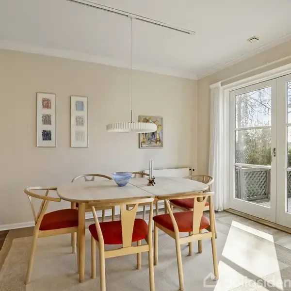 Et spisebord med seks røde stole står i et lyst rum. En moderne lampe hænger over bordet. Vinduerne til højre slipper lys ind og viser en have udenfor.