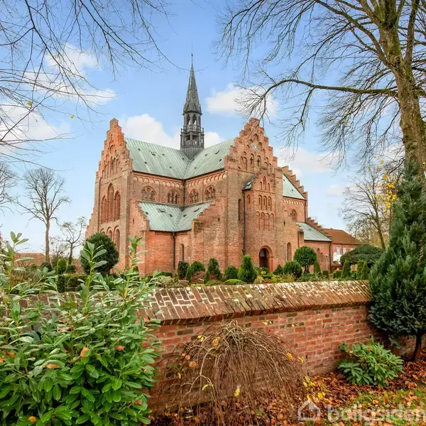 Kirkebygning står majestætisk, omgivet af grønne buske og træer, med en blå himmel i baggrunden. Foran ses en mur af røde mursten, der indrammer stedet.