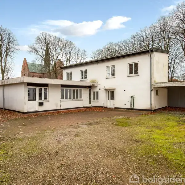 Hvid bygning med fladt tag står stille i en åben gård omkranset af bare træer og en grusbelagt gårdsplads.