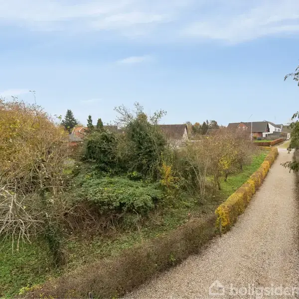 Grusvej løber gennem en have med buske på begge sider. Omkringliggende træer og buske skaber en naturlig ramme. I baggrunden ses et boligområde med huse under en blå himmel.