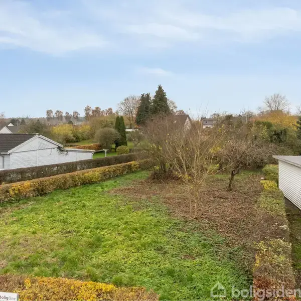 En græsplæne ligger bag en hæk i en boligkvarter. Til højre ses et hvidt hus, omgivet af buske og træer. Skyfri blå himmel.
