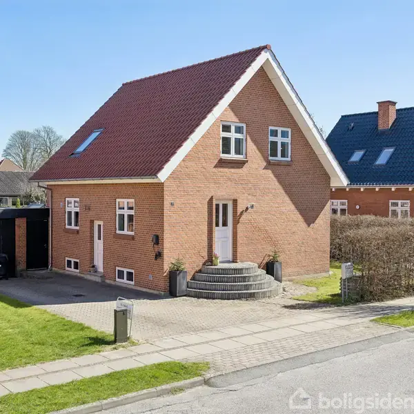 Rødstenshus med rødt tag har en trappe foran hoveddøren. Til venstre ses en garage med sort tag. Omgivet af grønne græsplæner og en hæk, står huset i et beboelseskvarter.