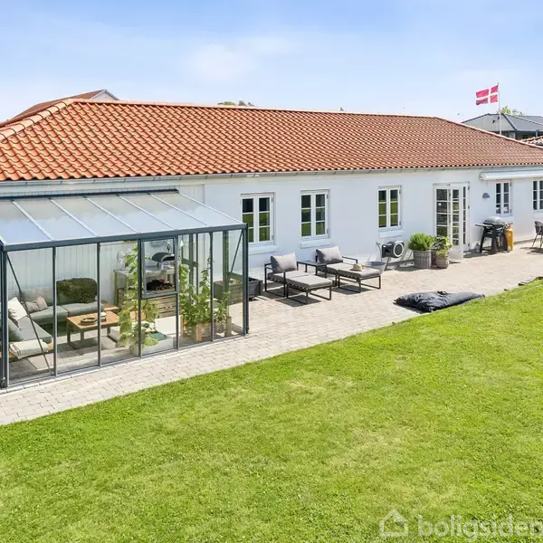 Hvidt hus med rødt tegltag, har en tilstødende udestue med siddemøbler. Terrasseområdet har bord og stole, omkranset af velplejet græsplæne. En dansk flag er hejst i baggrunden.