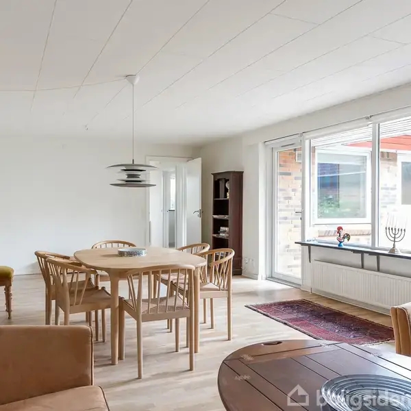 Spisebord omgivet af stole står i en lys dagligstue med hvide vægge. Store vinduer til højre giver udsigt til en terrasse. En reol og skænk står i baggrunden. En brun sofa er synlig i forgrunden.