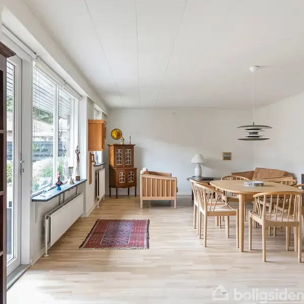 En stue med trægulv og spisebord med stole i centrum. Store vinduer til venstre giver udsigt til haven. Reoler og kommoder med dekorative genstande står langs væggene.