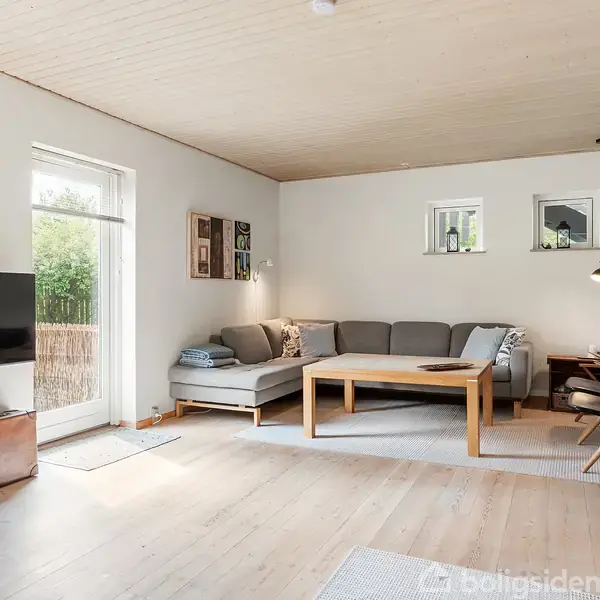 Stue med en grå hjørnesofa og træbord i centrum. TV på væggen til venstre. Trægulv og -loft. Store vinduer giver lys; indretningen er minimalistisk. Ingen tekst.
