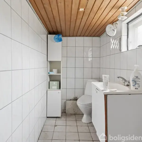 Et badeværelse med hvidt flisegulv og vægge. Et skab står i hjørnet ved siden af et toilet. En vask med sæbe og tandpasta er til højre under et vindue.