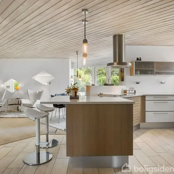 Køkkenø med høje stole omgivet af moderne køkkenudstyr, avantgarde lofthængte lamper og åbent rum, hvor en stue med sofagruppe og store vinduer ses i baggrunden.