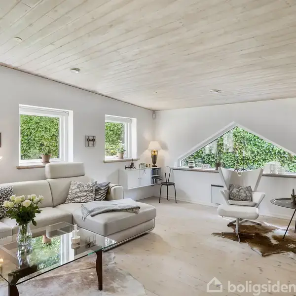 En lys stue med en hvid hjørnesofa, glasbord med blomster, og moderne stole ved et tæppe. Store vinduer lader naturligt lys ind, og udsigten er mod en grøn hæk.