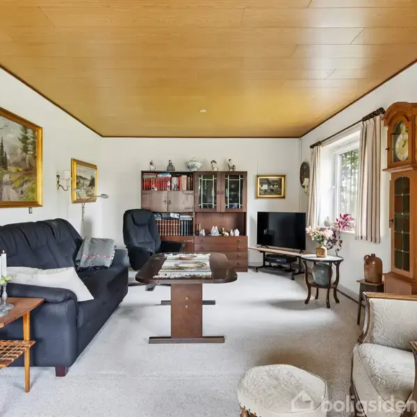 Stue med sort sofa og lænestol ved et sofabord. Reol med bøger og TV i baggrunden. Store vinduer med gardiner, og et ur står i hjørnet. Ingen tekst.