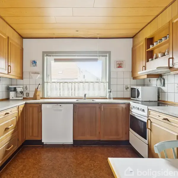 Køkken med træskabe og hvide fliser, udstyret med komfur, opvaskemaskine og mikrobølgeovn; vindue centralt i rummet med udsigt til udendørs område, placeret over vasken.