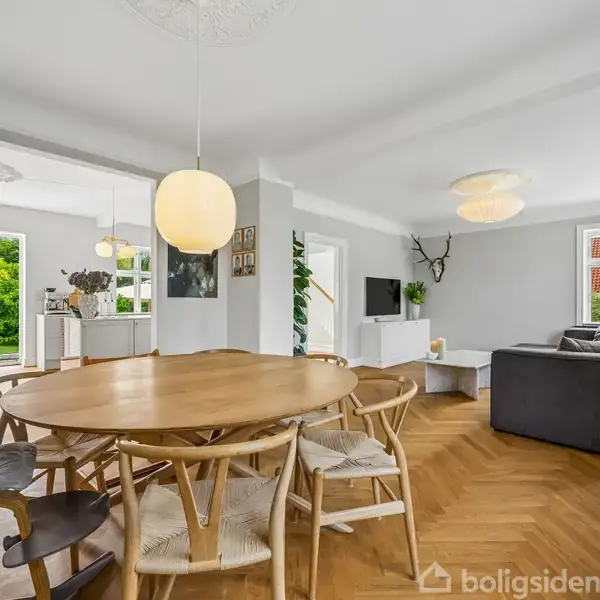 Rundt træbord omgivet af stole, placeret i en moderne stue med parketgulv, sofagruppe, og store vinduer mod haven. Lysekrone hænger over bordet, og en åben dør fører ud til en grøn have.