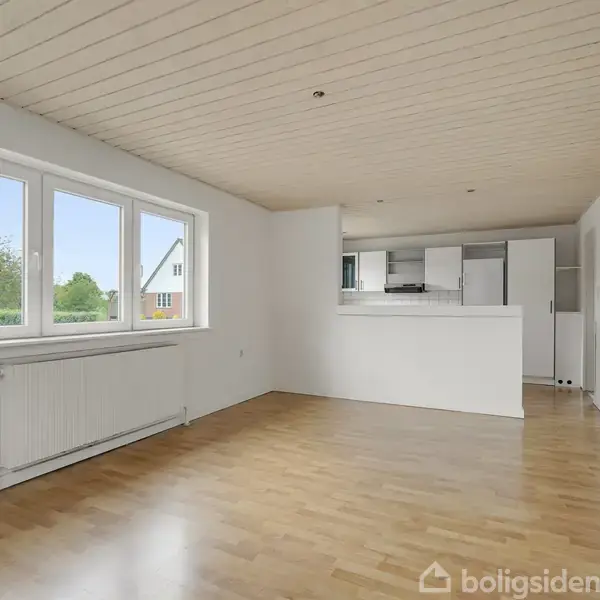 Stue med lyst trægulv og hvide vægge har store vinduer med udsigt til haven. I baggrunden ses et åbent køkken med hvide skabe og en hvid loftsbeklædning.