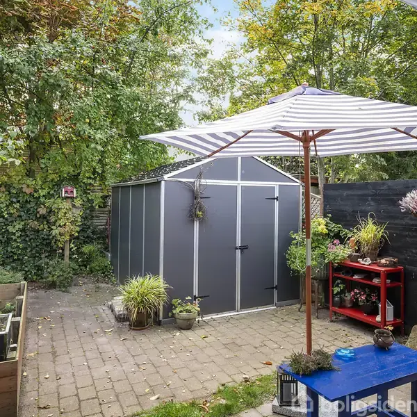 En have med et gråt skur står under en stribet parasol. En plantekasse og et rødt bord med planter omgiver området. Træer og buske danner en grøn baggrund.