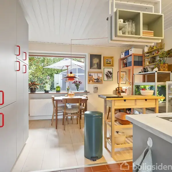 Et lyst køkken med grå skabe og røde håndtag står i forgrunden. En lille spiseplads med et bord og stole ses ved vinduet, omgivet af planter og vægdekorationer.