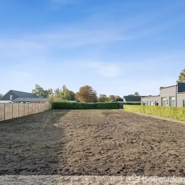 En bar jordgrund ligger stille mellem to hegn, omgivet af moderne huse og få træer under en klar blå himmel.