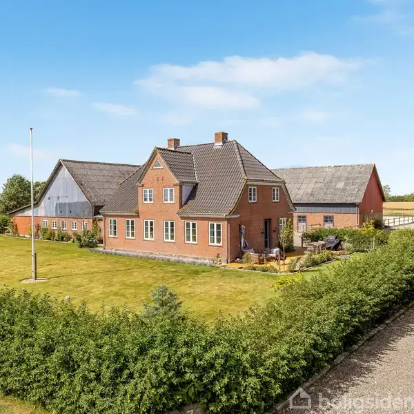 Murstenshus står stille omgivet af grønne hække og græsområde. Indkørsel til højre. Klar blå himmel i landligt område.