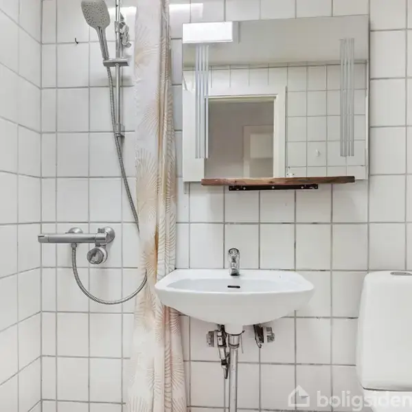 Et badeværelse med en bruser, siddende ved siden af en vask med spejl over, og et toilet ved siden af. Væggene er dækket af hvide fliser. Ingen tekst synlig.