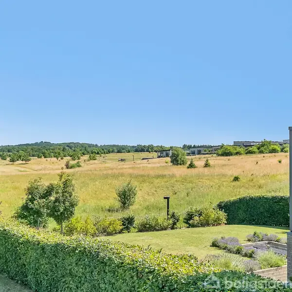 Hus med glasvæg står i rolig, landlig have med buske og velfriseret græsplæne; store marker og skov ses i det fjerne under en klar blå himmel.