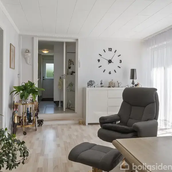 En komfortabel lænestol står i en lys stue med en sofabord og planter omkring sig. Et stort ur hænger på væggen over en kommode med forskellige dekorationer.