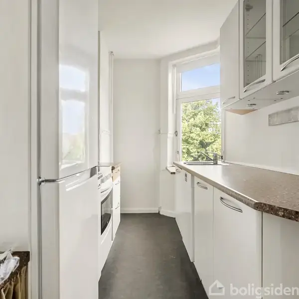Et smalt køkken med hvide skabe og mørk bordplade fører til et vindue, der lukker lys ind. Den lyse atmosfære understreges af minimalistisk indretning og en grøn udsigt udenfor.