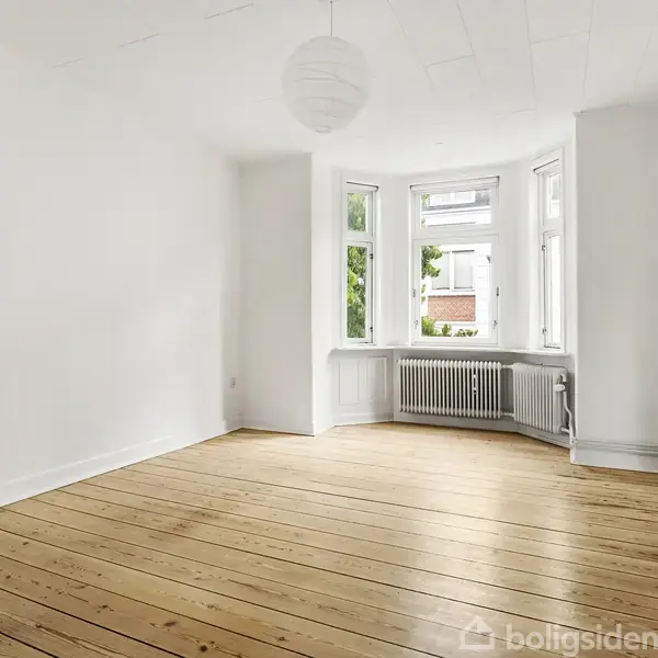 Et tomt værelse har lyse trægulve og hvide vægge. En stor vinduesbue giver udsigt til træer udenfor. En radiator er placeret under vinduet, og en rund loftslampe hænger centralt.