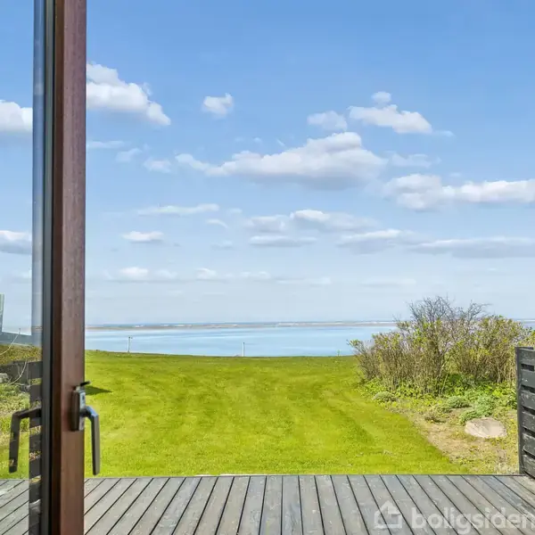 Åben dør fører ud til en træterrasse, der strækker sig mod en velplejet græsplæne, med en betagende udsigt over havet i baggrunden under en klar blå himmel.
