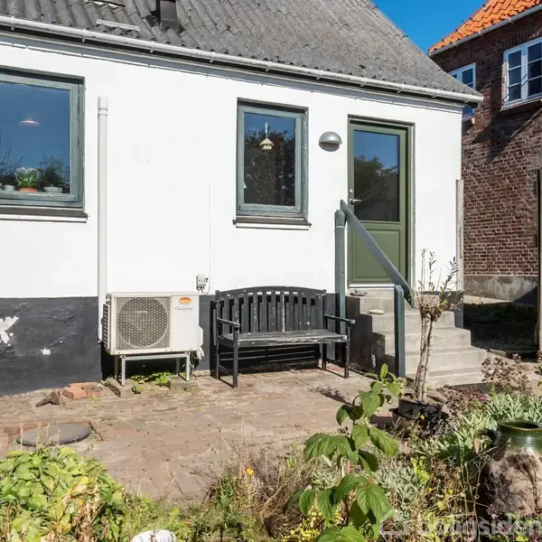 En hvidmalet baggårdsmur med to vinduer og en dør, en sort bænk placeret foran. Trappen fører til en lille, blomsterfyldt gårdhave.