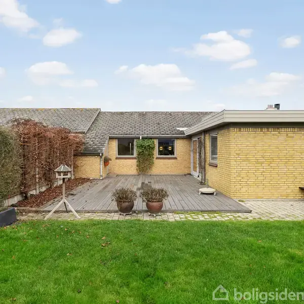 Et gult murstenshus med fladt tag står stille. En træterrasse med potteplanter og fuglefoderstation ses i forgrunden, omgivet af grøn græsplæne og buske under en blå himmel.