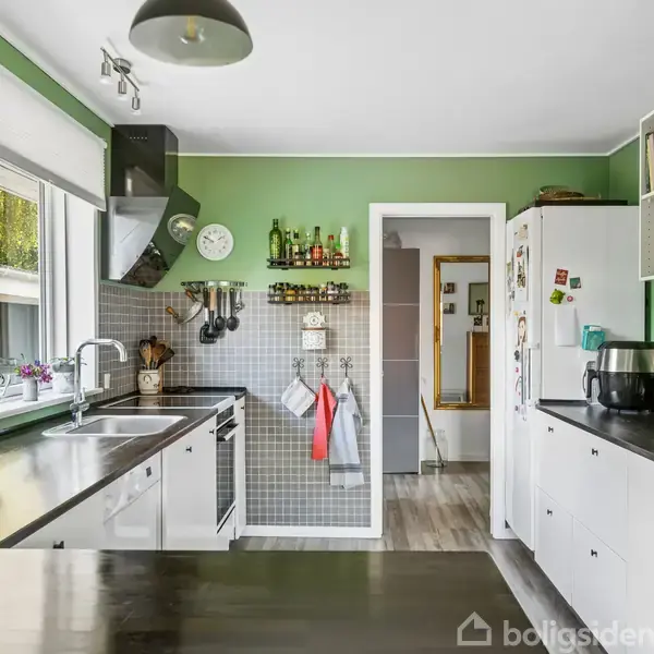 Køkken med grønne vægge indeholder køkkenbord, hvide skabe, og apparater som mikrobølgeovn og køleskab. Et vindue med potteplanter giver lys. Tekst på tavlen: "Kiss the cook."
