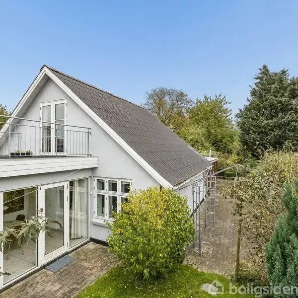 Et hvidt hus med gavltag står stille i en frodig have. Store glasdøre åbner ud mod en terrasse. Omkringliggende træer og buske skaber en grøn atmosfære.