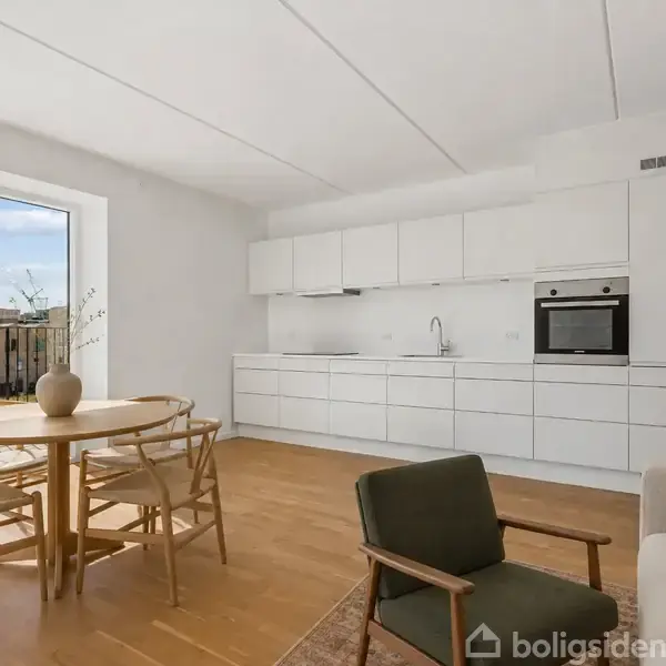 Et moderne køkken-alrum med et rundt træbord og fire stole. Køkkenet har hvide skabe og en indbygget ovn. Store vinduer giver udsigt til en terrasse og bygninger udenfor.