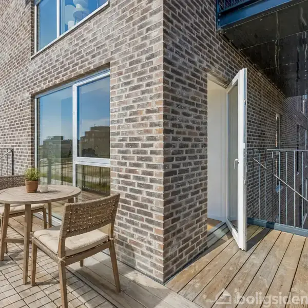 En træterrasse med et rundt bord og to stole står langs en murstensbygning. En åben dør fører ind i lejligheden. Planter og glasvinduer omkranser stedet.