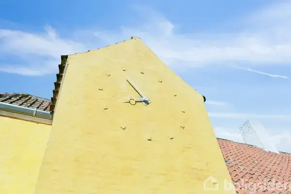 En stor, gul murvæg har et ur uden cifre. Viserne står stille. Omkring er der røde tegltag og en klar blå himmel med få skyer.