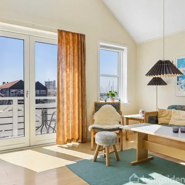 En stue med et lille bord og bænk, oplyst af solskin gennem glasdøre med orange gardiner, har en hyggelig atmosfære. Der hænger en pendellampe og billeder på væggen.