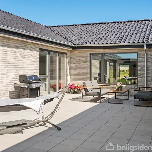 En åben gårdhave med fliser, der har en hængekøje og grill på den ene side. To moderne stole og et bord står i midten. Murstenshus omgiver gårdhaven.