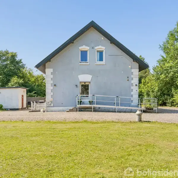 Et gråt hus med mørkt tag har en rampe ved indgangen. Huset står på en grusvej omgivet af træer og en grøn græsplæne.
