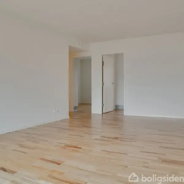 Et tomt rum med lyst trægulv strækker sig mod en åben dør. Væggene er hvide, og rummet har en minimalistisk atmosfære i et boligmiljø.