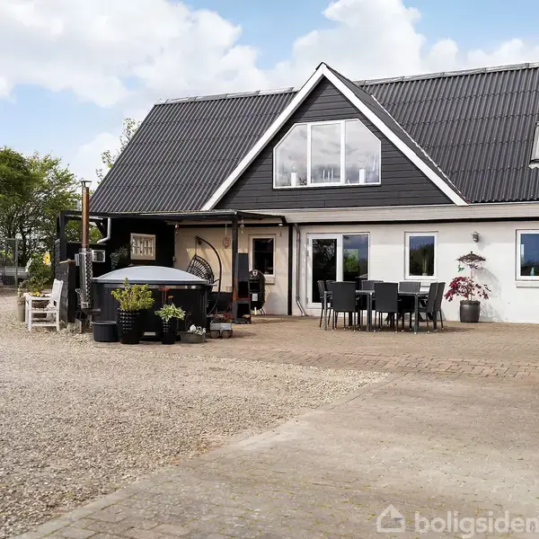 Et moderne hus med sort tag og stor glasgavl. I forgrunden er der havemøbler og planter på en flisebelagt terrasseomgivet af grusvej, omkranset af træer.