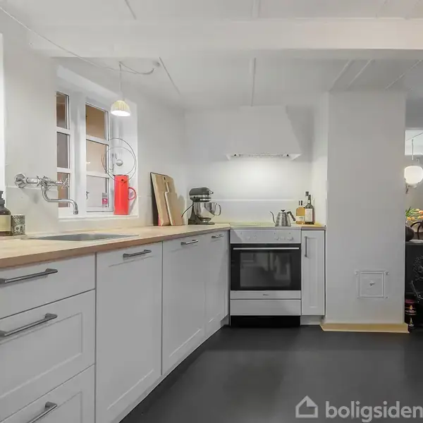 Køkken med hvide skabe og træbordplade, stående ovn, placeret i et lyst rum med sorte gulve. Vinduet over vasken slipper lys ind, og der er moderne køkkenudstyr.