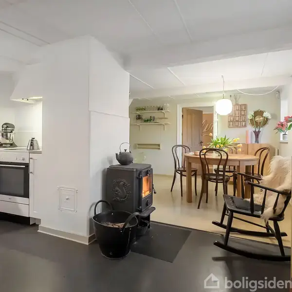 Brændeovn opvarmer stue-køkkenområde; køkken med komfur til venstre, spisebord med stole i baggrunden, og en gyngestol til højre. Hyggelig atmosfære med planter og lyse farver.