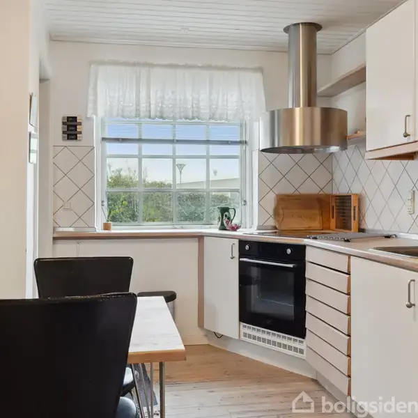 Et køkken med spisebord og stole står ved siden af et vindue. Bordet har en plante som dekoration. Komfuret og vasken er indbygget i en hvid indretning, omkranset af trædetaljer.